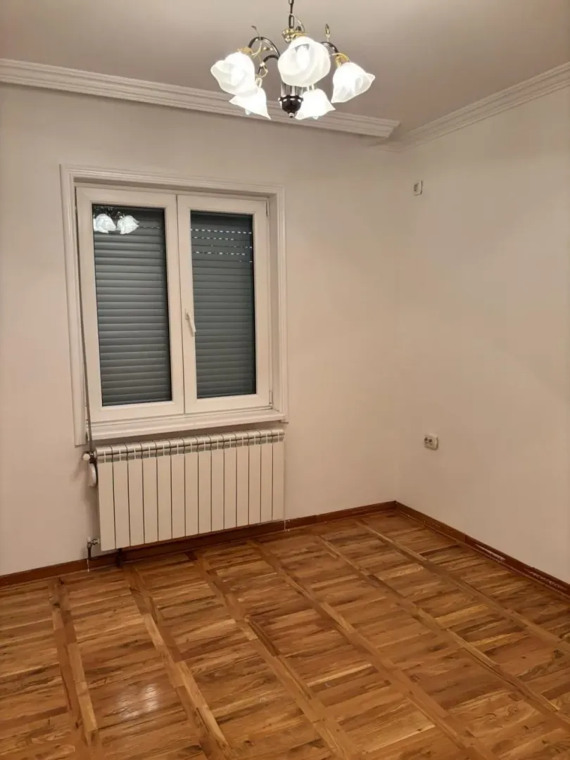 Empty room with hardwood floors, a window with roller blinds, and a ceiling chandelier.