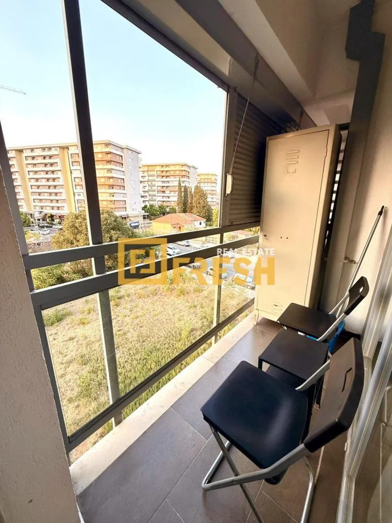 Zastakljeni balkon sa stolicama, ormarićem i pogledom na okolne zgrade.