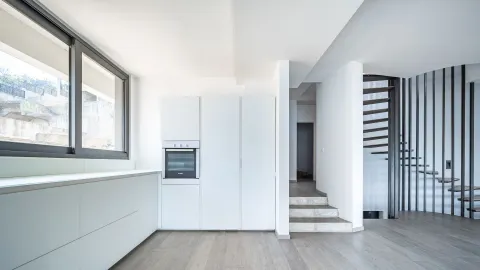 Moderna bijela kuhinja sa ugrađenom rernom pored stepeništa i velikim prozorom. - image 13