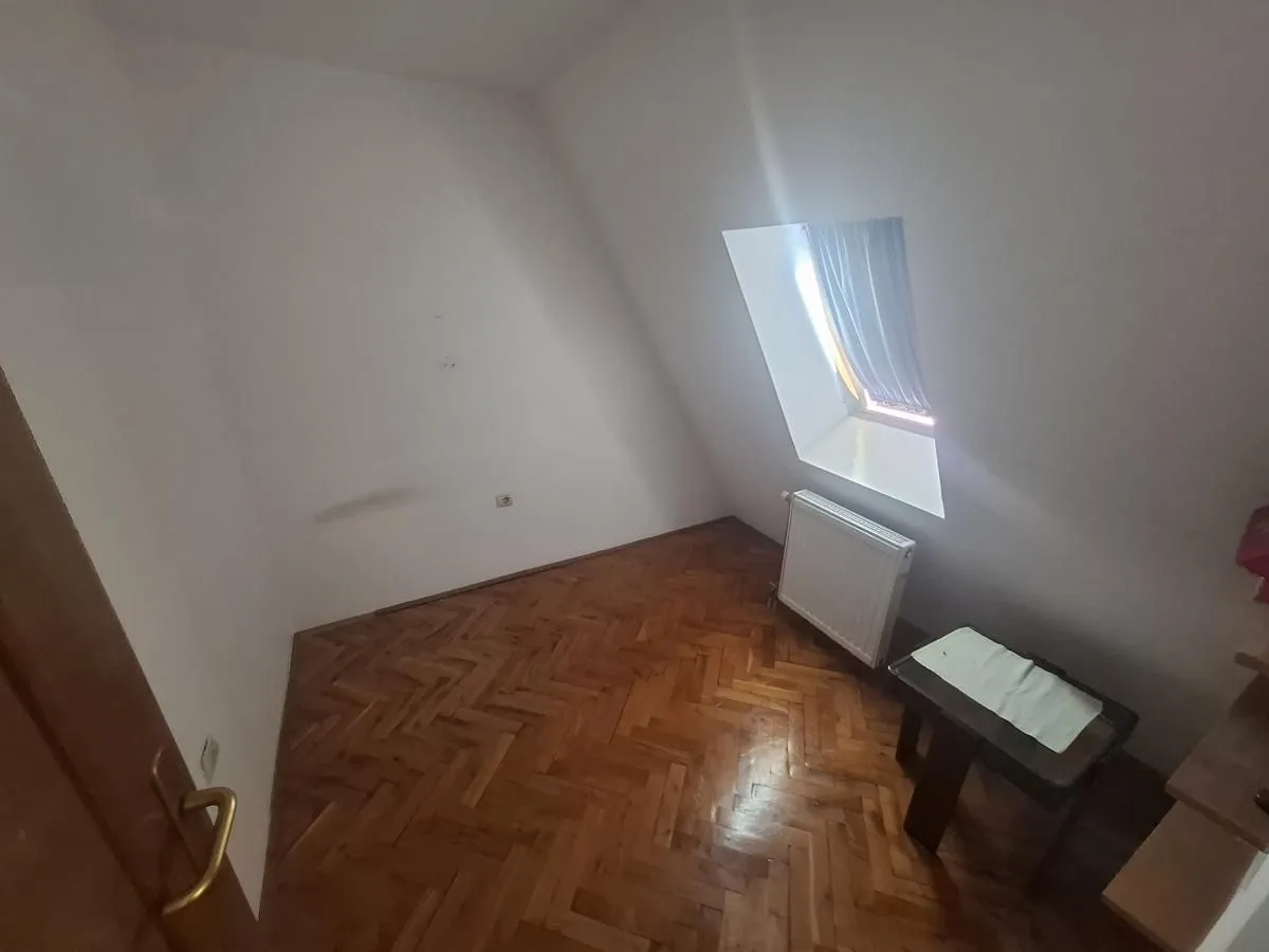 Prazna soba sa kosim krovom, parketom i malim prozorom.