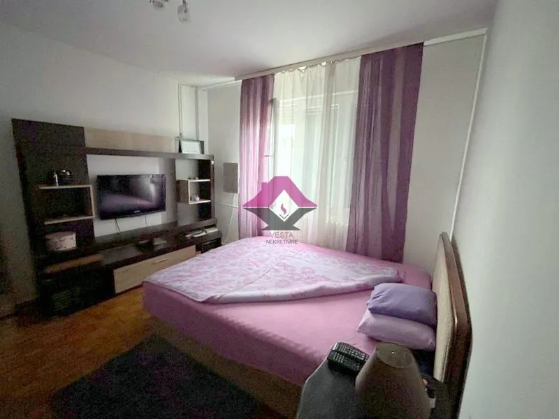 Nameštena spavaća soba sa bračnim krevetom, TV-om i prozorom sa ljubičastim zavesama.