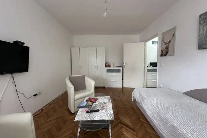 Nameštena spavaća soba sa krevetom, foteljom, ormanom i televizorom na zidu.