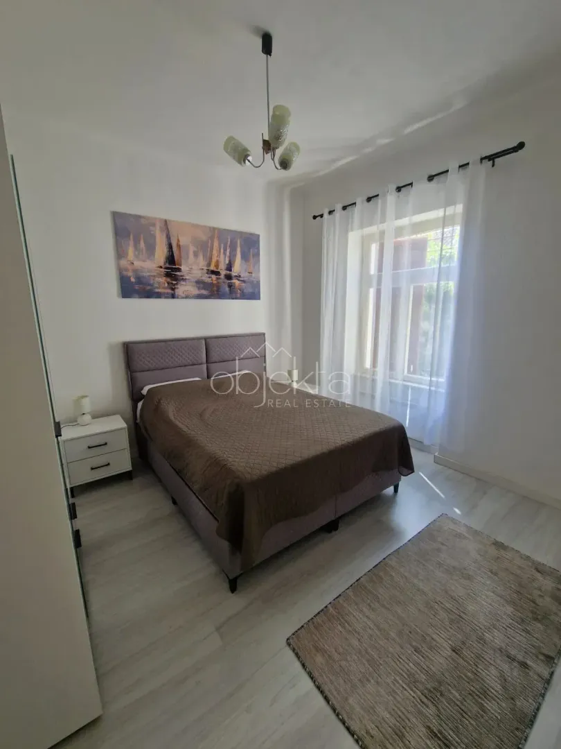 Spavaća soba sa bračnim krevetom, noćnim stočićem i prozorom sa bijelim zavjesama.
