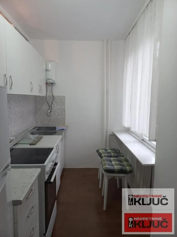 Manja kuhinja sa belim elementima i šankom sa stolicama pored prozora.