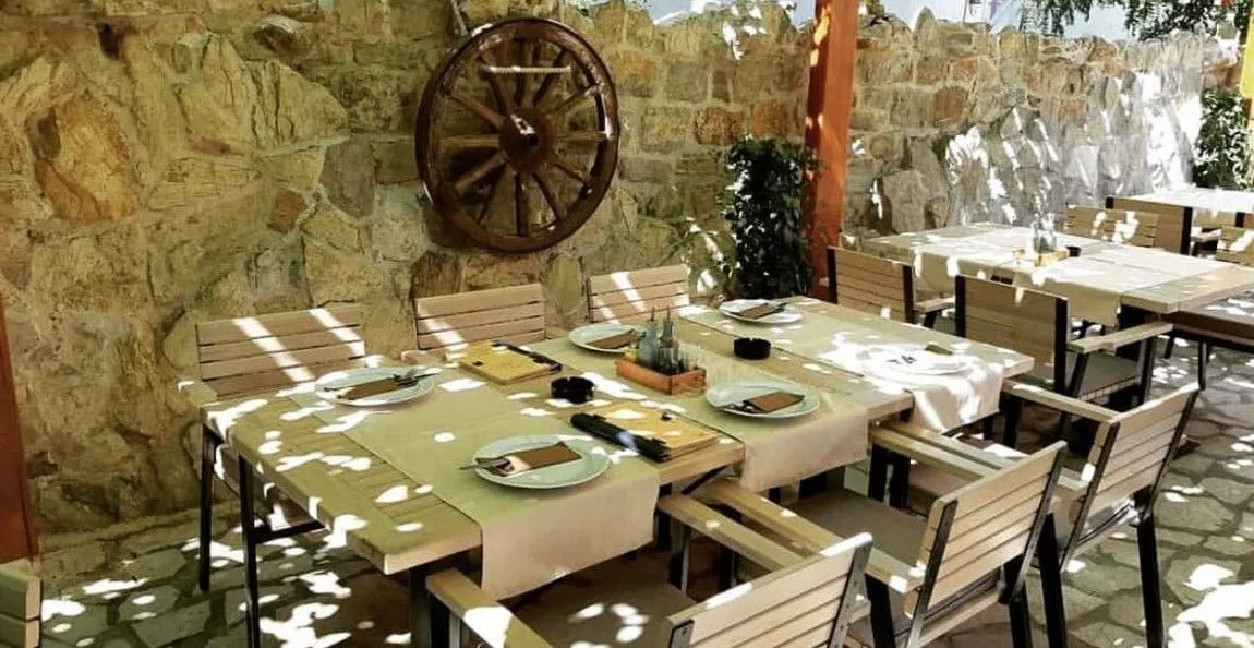 Terrace with wooden tables and chairs next to a stone wall decorated with an old wheel.
