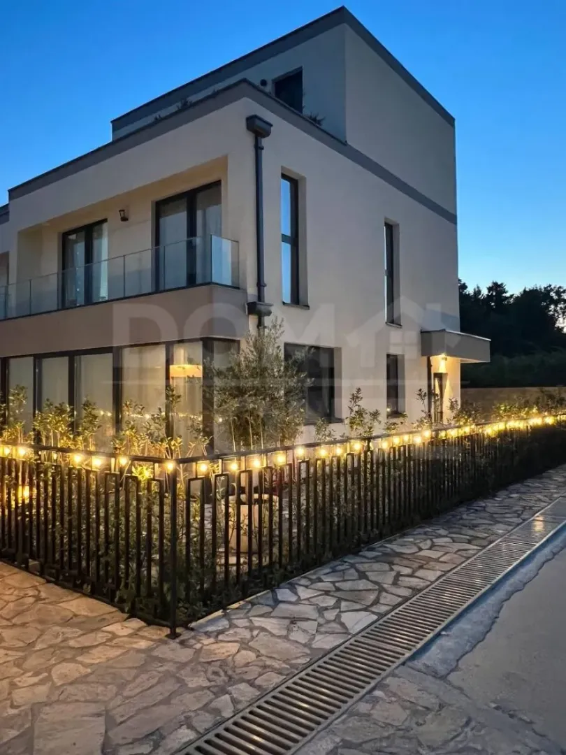 Exterior of a modern house with a yard and illuminated fence at dusk.