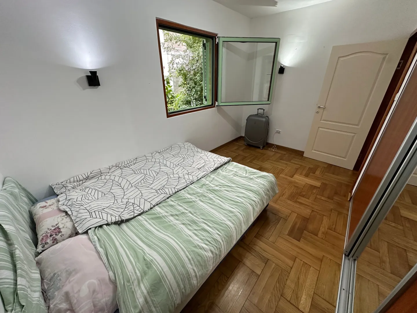 A bedroom with a bed, wooden flooring, and a window overlooking greenery.