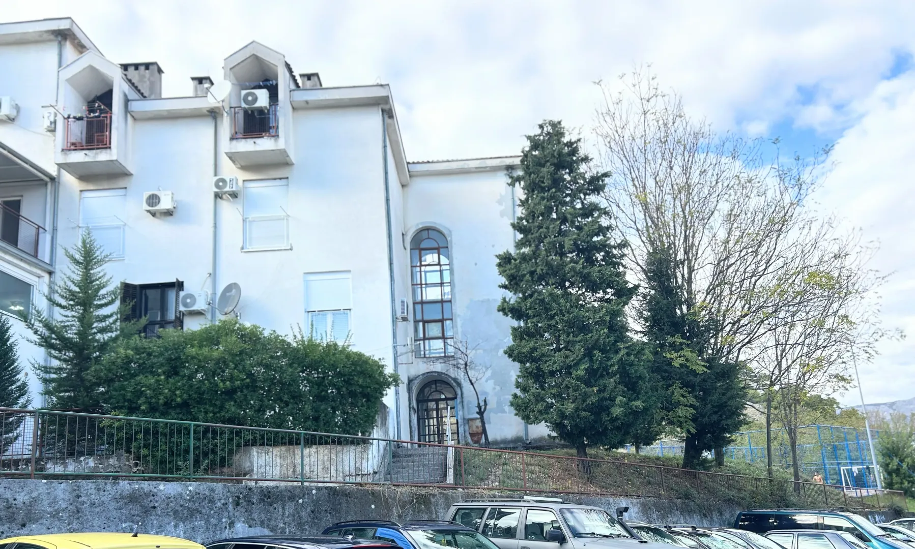 Spoljašnji izgled bijele stambene zgrade sa dvorištem i parkiranim automobilima.