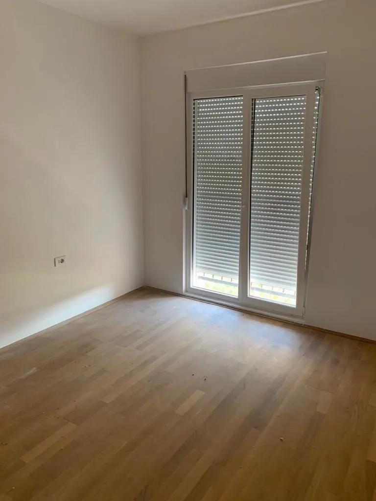 Prazna soba sa drvenim podom i balkonskim vratima sa roletnama.