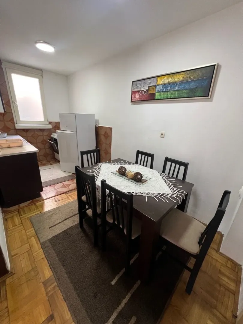 Trpezarija povezana sa kuhinjom sa bijelom tehnikom i drvenim stolom.