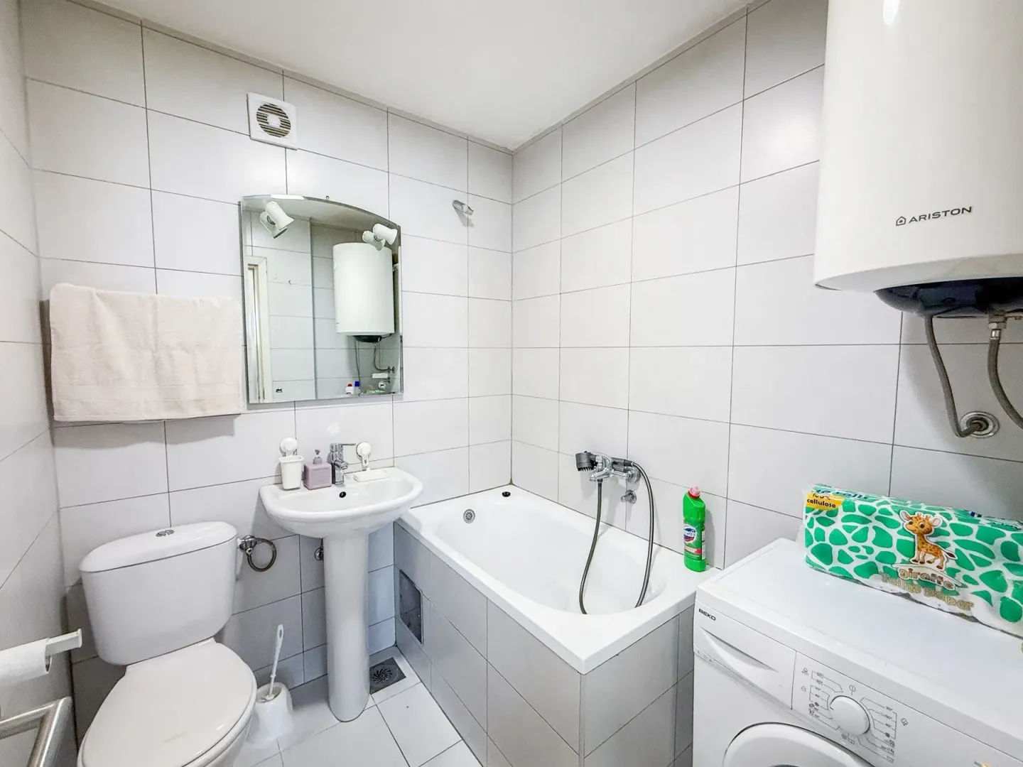 Small bathroom tiled in white with a bathtub, washbasin, toilet, and washing machine.