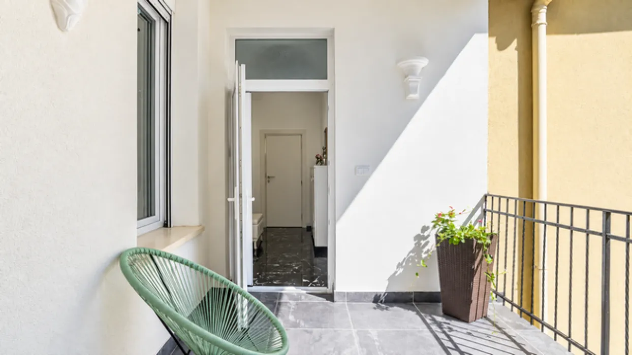 Manji svetli balkon sa metalnom ogradom, stolicom i saksijom sa cvećem.