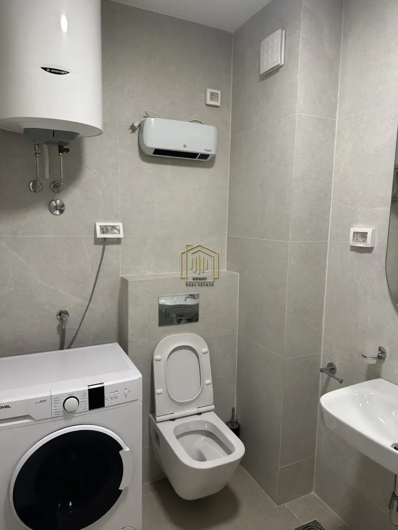 Bathroom tiled in grey with a toilet, washbasin, and washing machine.
