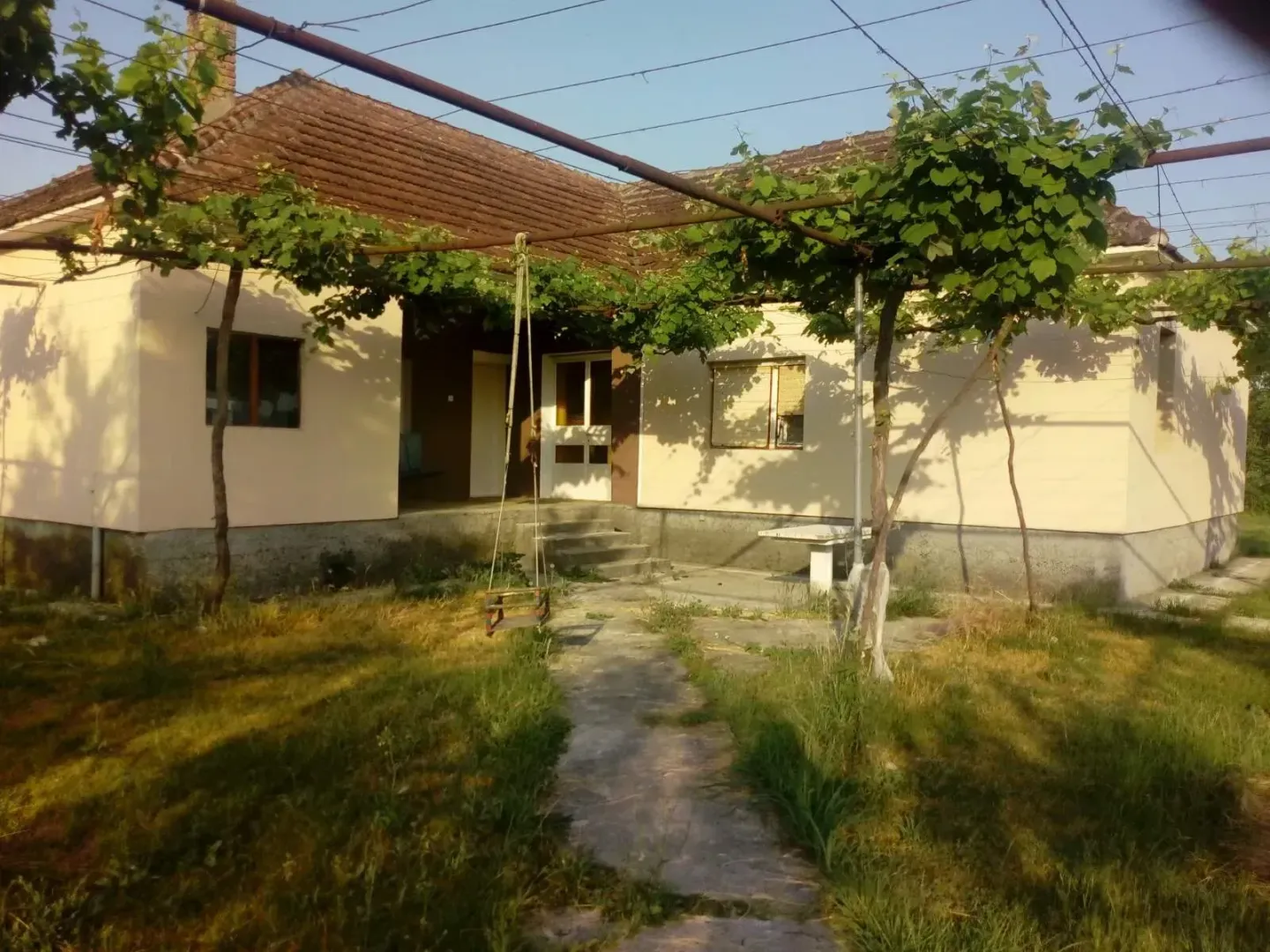 Pogled na dvorište i fasadu kuće sa braon krovom i natkrivenom lozom.