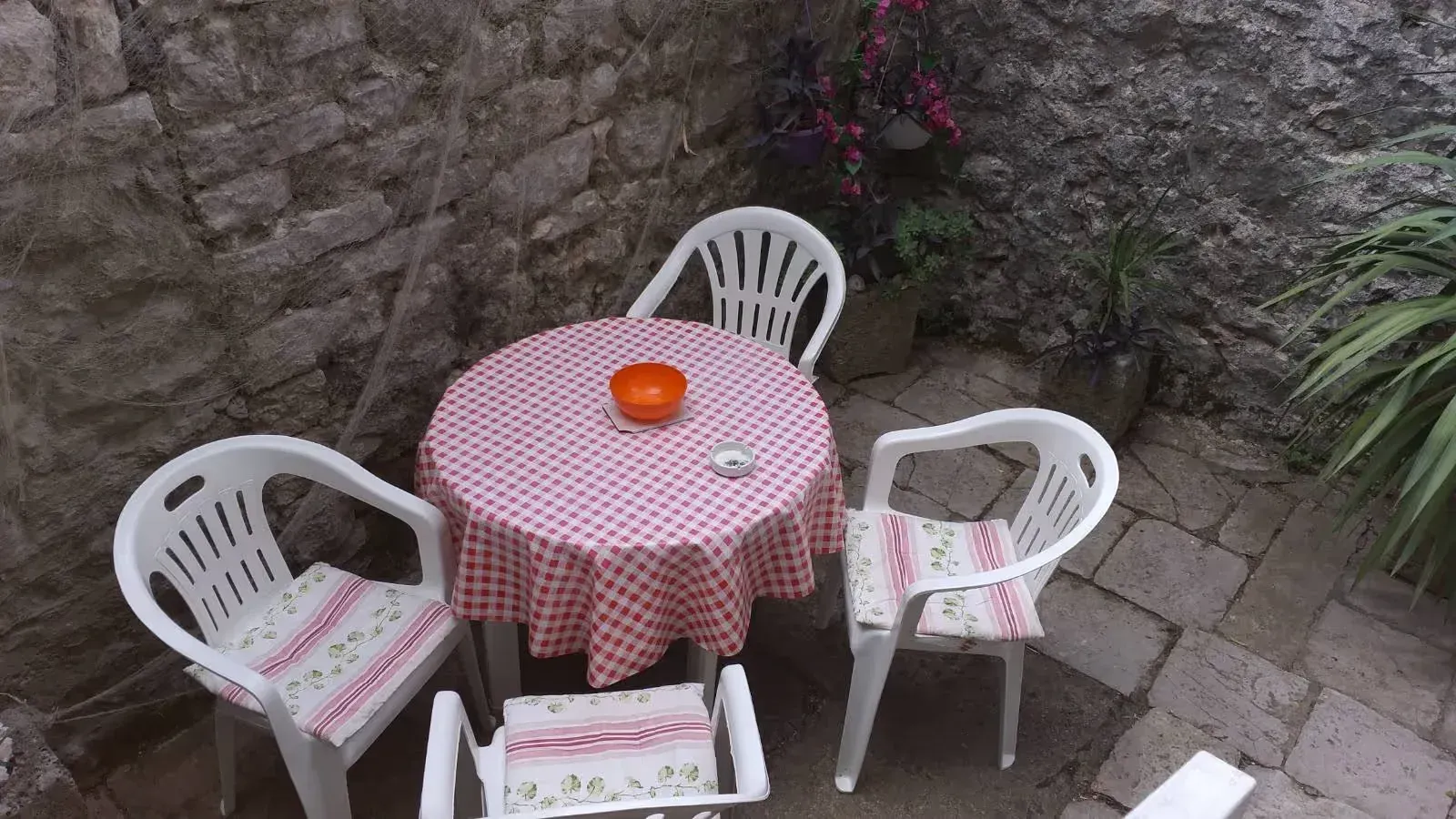 Mali popločani dvorišni prostor sa stolom i bijelim plastičnim stolicama pored kamenog zida.