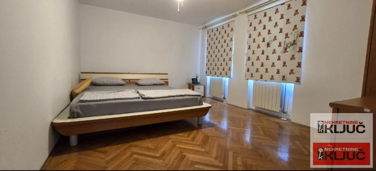 Bedroom with a double bed, wooden parquet flooring, and windows with patterned roller blinds.