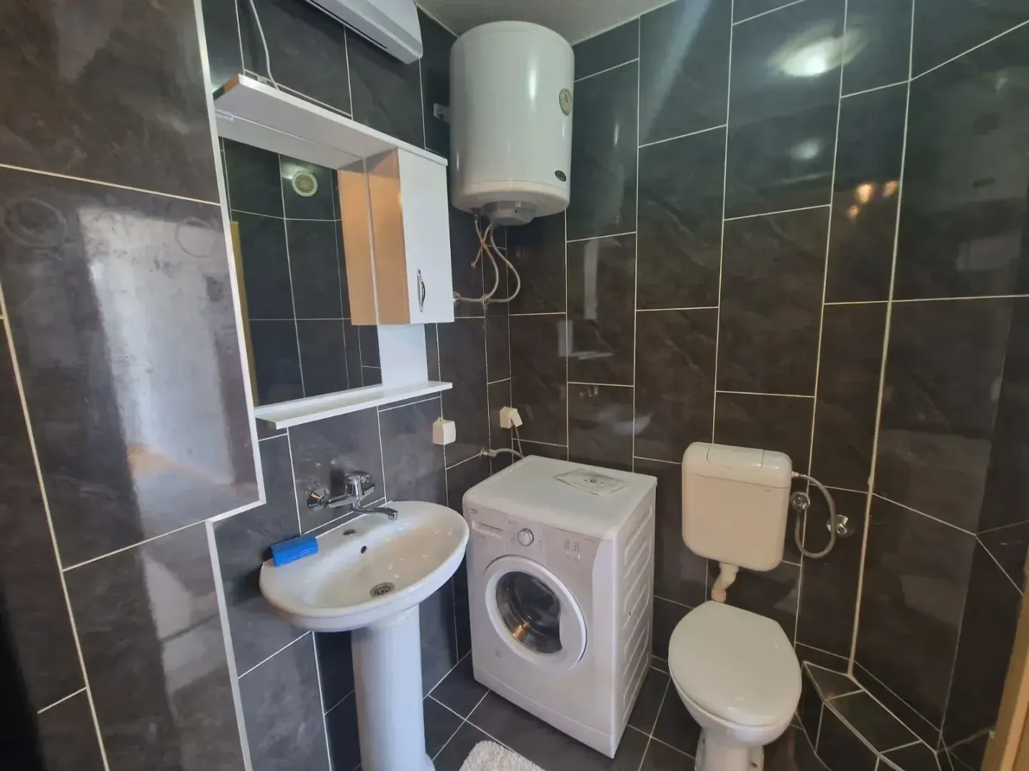 Bathroom equipped with a sink, washing machine, toilet, and dark tiles.