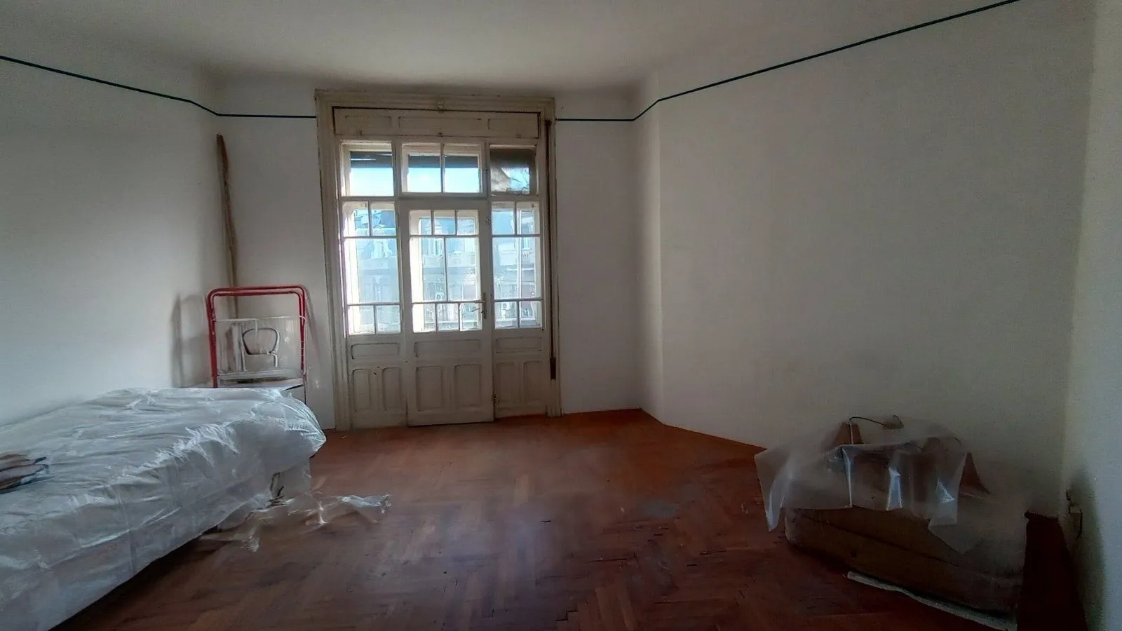 Prazna soba sa drvenim podom, velikim dvokrilnim vratima i krevetom prekrivenim zaštitnom folijom.
