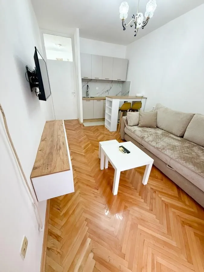 Dnevna soba sa garniturom, televizorom i kuhinjom u pozadini.