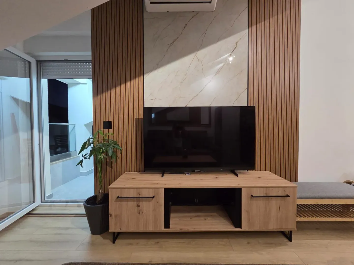 Dnevna soba sa TV komodom, drvenim zidnim panelima i izlazom na terasu.