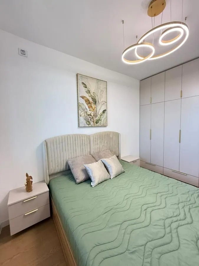 Nameštena spavaća soba sa bračnim krevetom, ormarom i modernim lusterom.