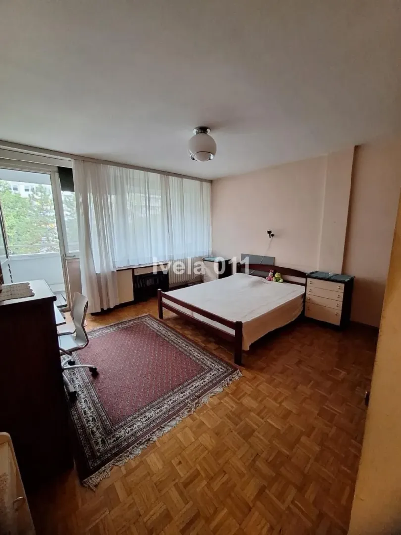 Nameštena spavaća soba sa bračnim krevetom, radnim stolom i izlazom na terasu.