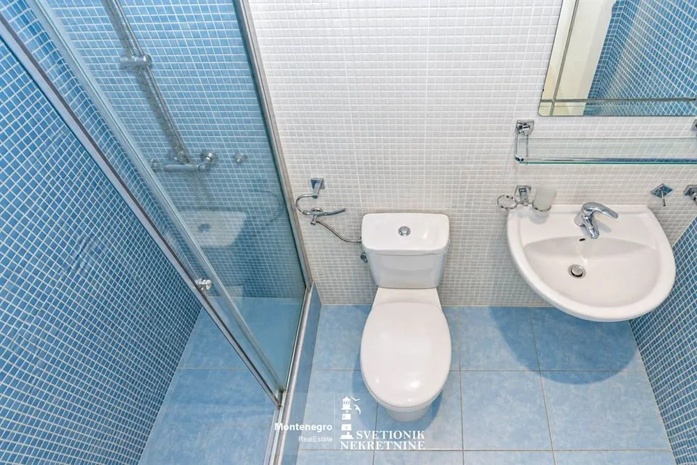 Manje kupatilo obloženo plavim pločicama sa tuš kabinom, WC šoljom i umivaonikom.