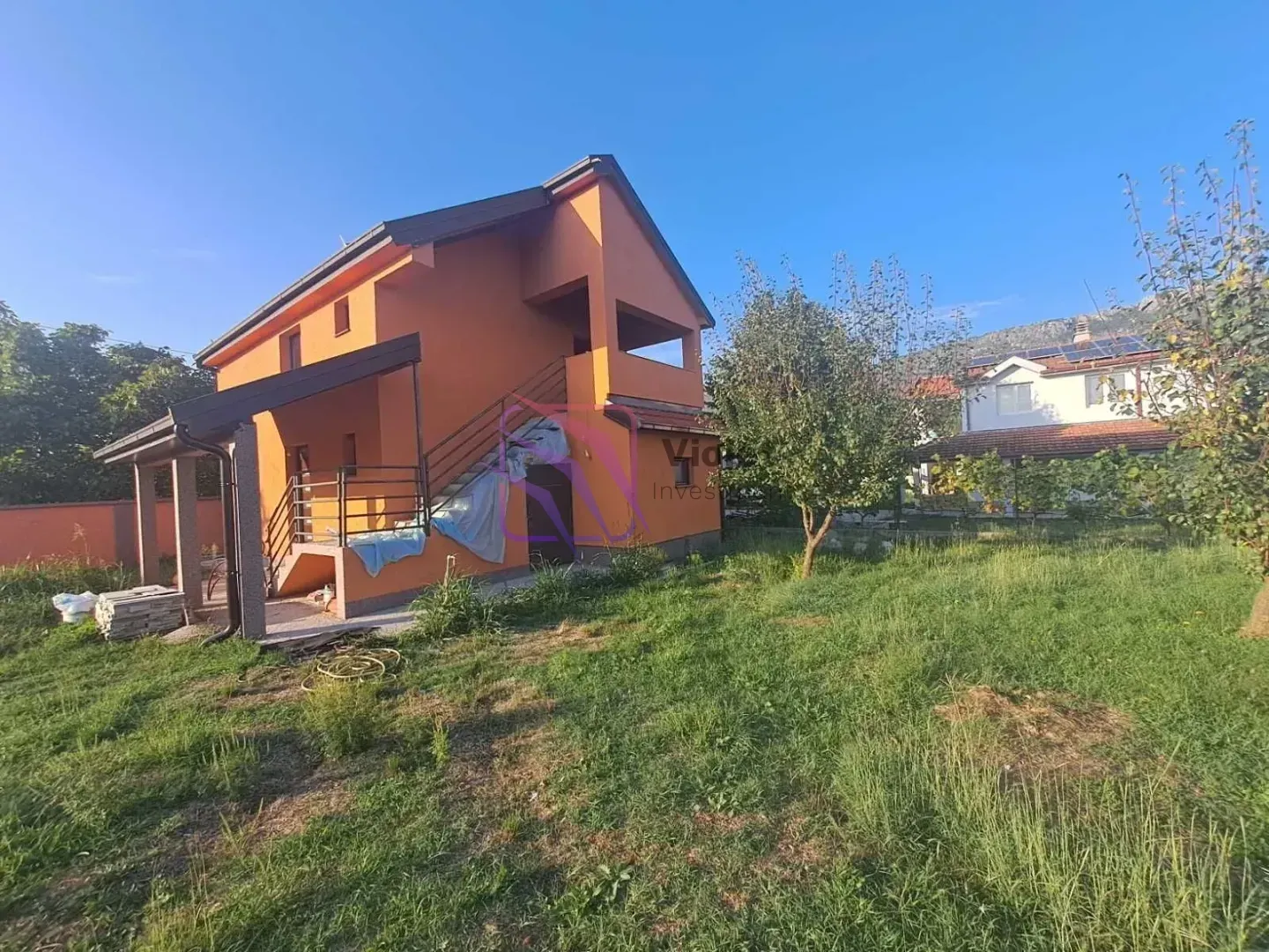 Pogled na dvorište i fasadu dvospratne kuće narandžaste boje sa spoljnim stepeništem.