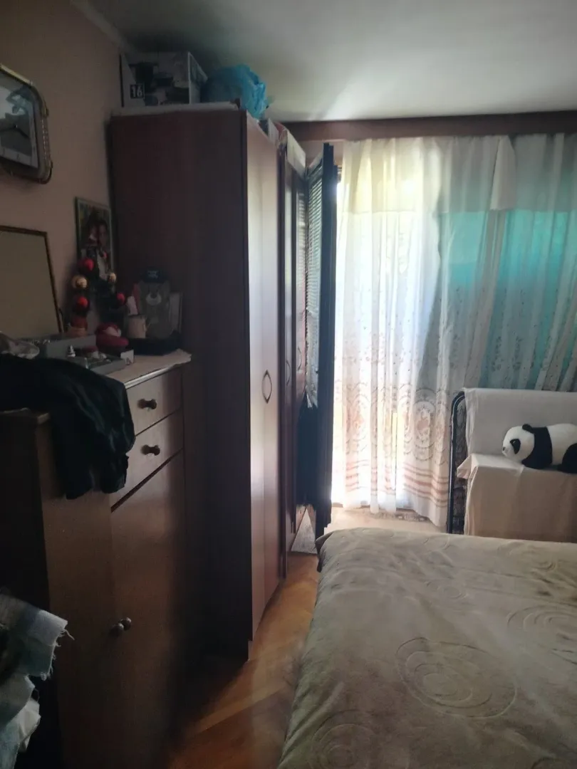 Spavaća soba sa velikim ormarom, komodom i krevetom pored prozora sa zavjesama.