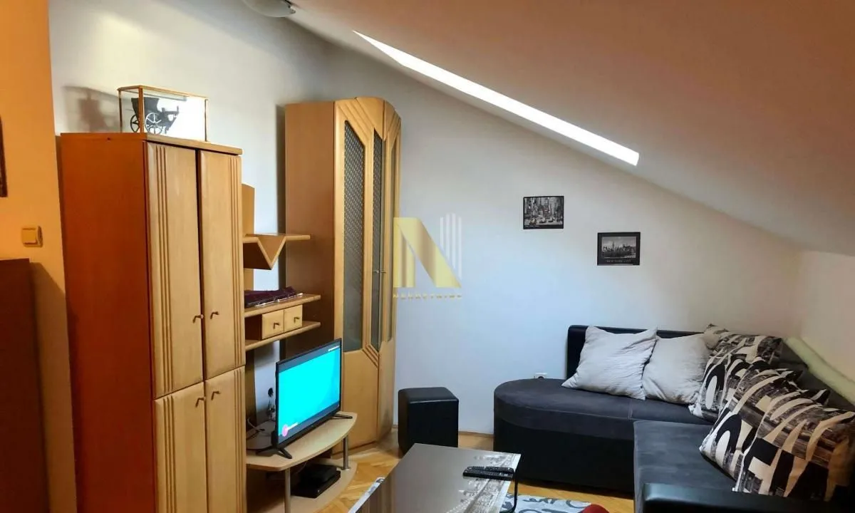 Dnevni boravak sa garniturom, televizorom i drvenim ormarima pod kosim krovom.