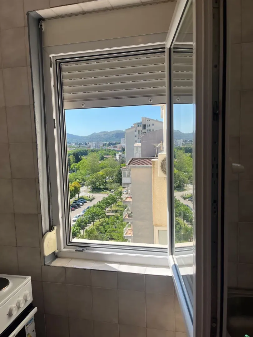 Pogled kroz otvoreni prozor na zelenilo i okolne zgrade.