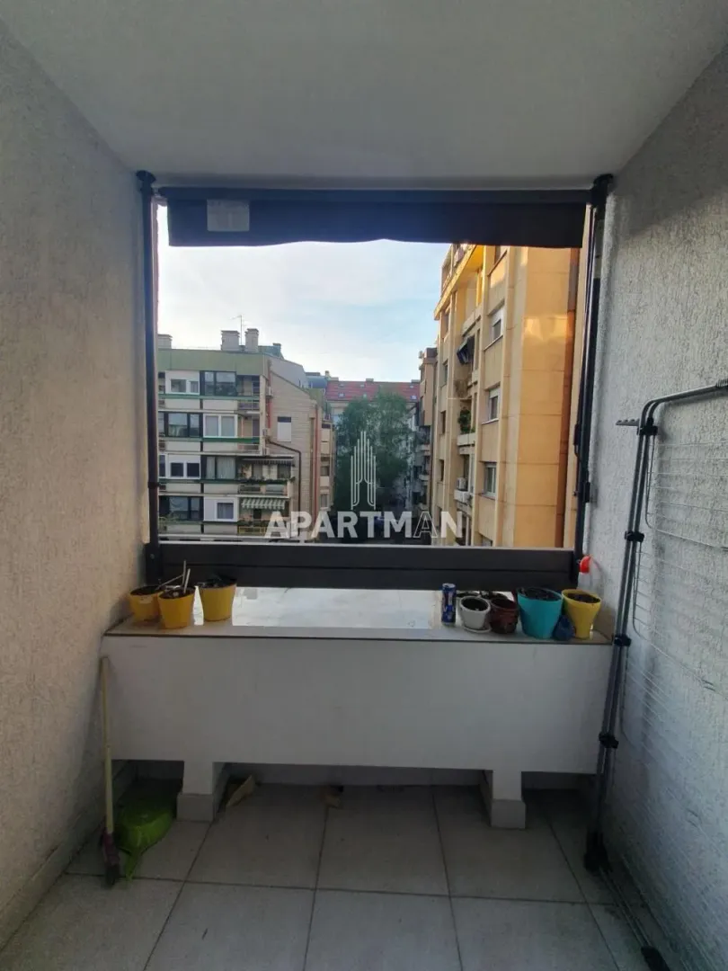 Manji zatvoreni balkon sa pogledom na okolne zgrade i saksijama sa cvećem.