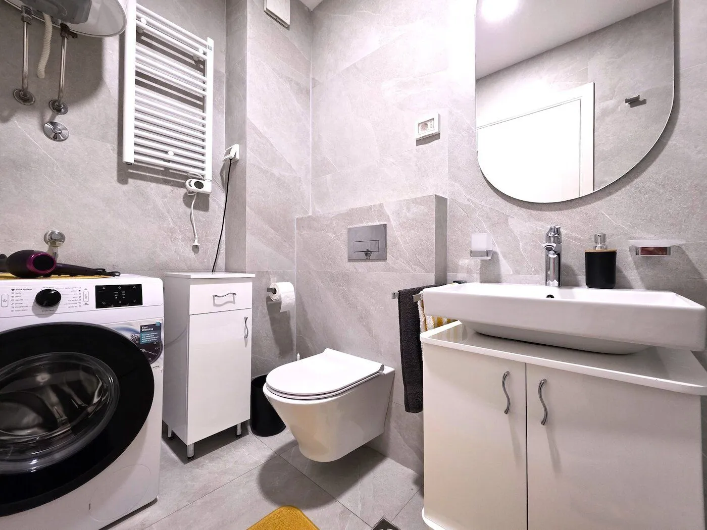 Modern bathroom with a washing machine, vanity unit, and toilet finished in grey tiles.