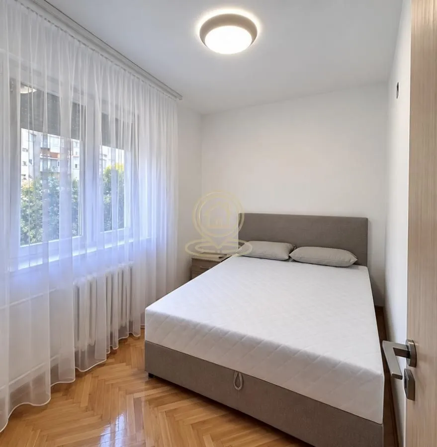 Manja spavaća soba sa bračnim krevetom, parketom i prozorom sa belim zavesama.