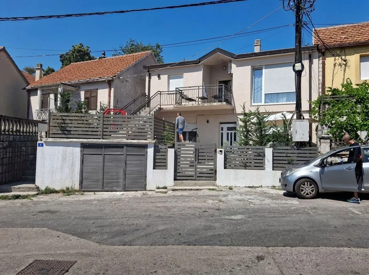 Pogled na spoljašnjost kuće sa dvorištem, ogradom i parkiranim automobilom.