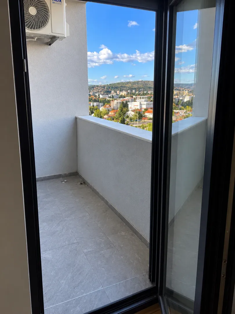 Pogled sa balkona na grad i okolna brda kroz otvorena balkonska vrata.