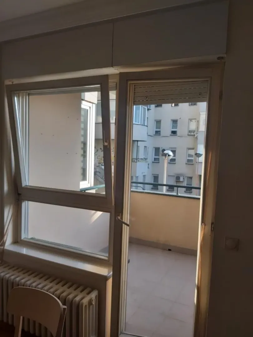 Pogled kroz balkonska vrata i prozor na spoljašnji deo i okolne zgrade.