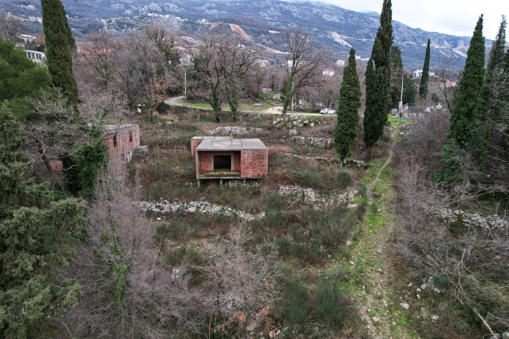 Nedovršeni objekat na prostranom placu okruženom drvećem i brdima.