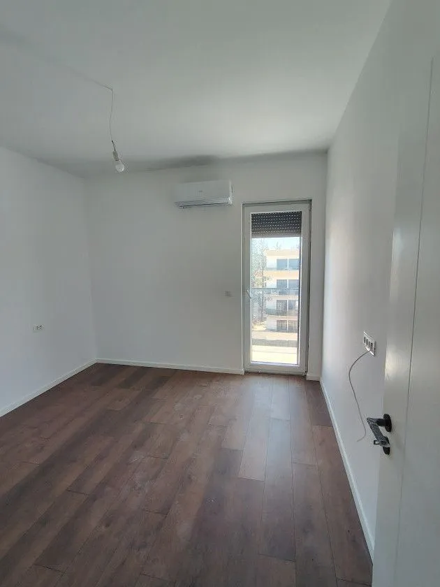 Prazna soba sa drvenim podom, klima uređajem i balkonskim vratima.