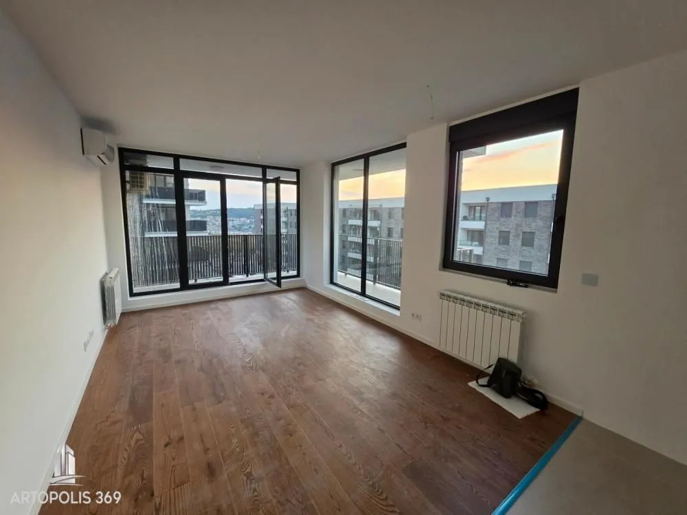 Empty living room with wooden flooring and large windows overlooking surrounding buildings.