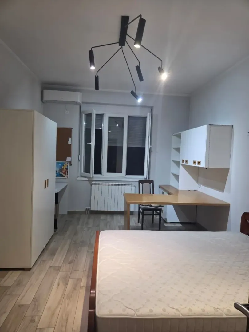 Namještena spavaća soba sa krevetom, radnim stolom, garderoberom i modernim lusterom.