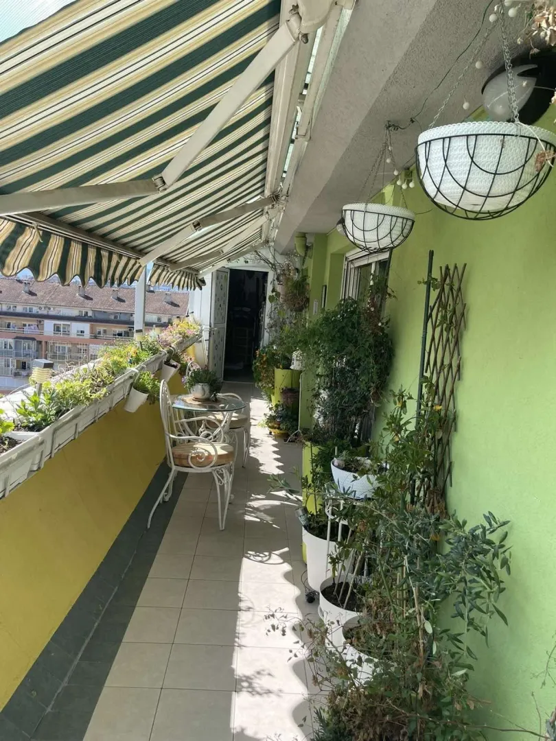Dugačak balkon sa garniturom za sjedenje i mnoštvom saksijskog cvijeća.