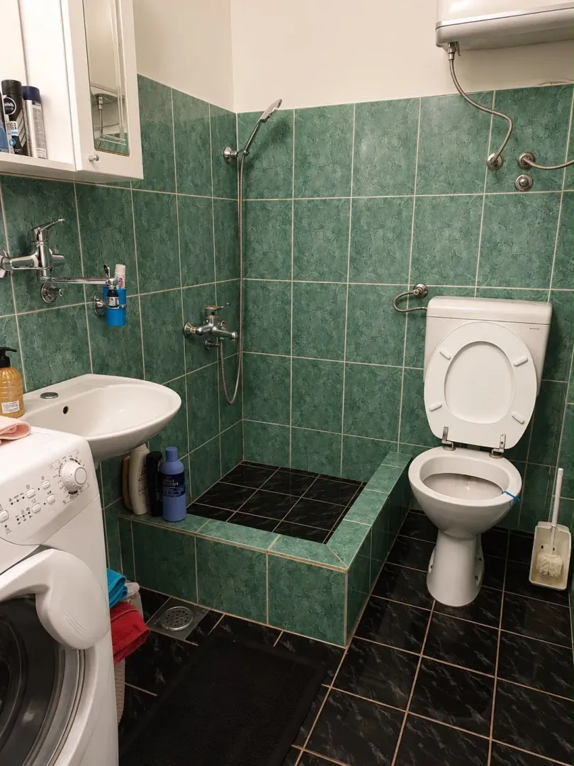 Manje kupatilo sa zelenim zidnim pločicama, tuš kabinom, umivaonikom i veš mašinom.