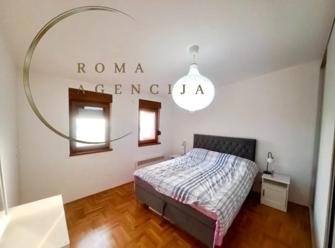 Bright bedroom with a double bed, wooden parquet flooring, and two windows.