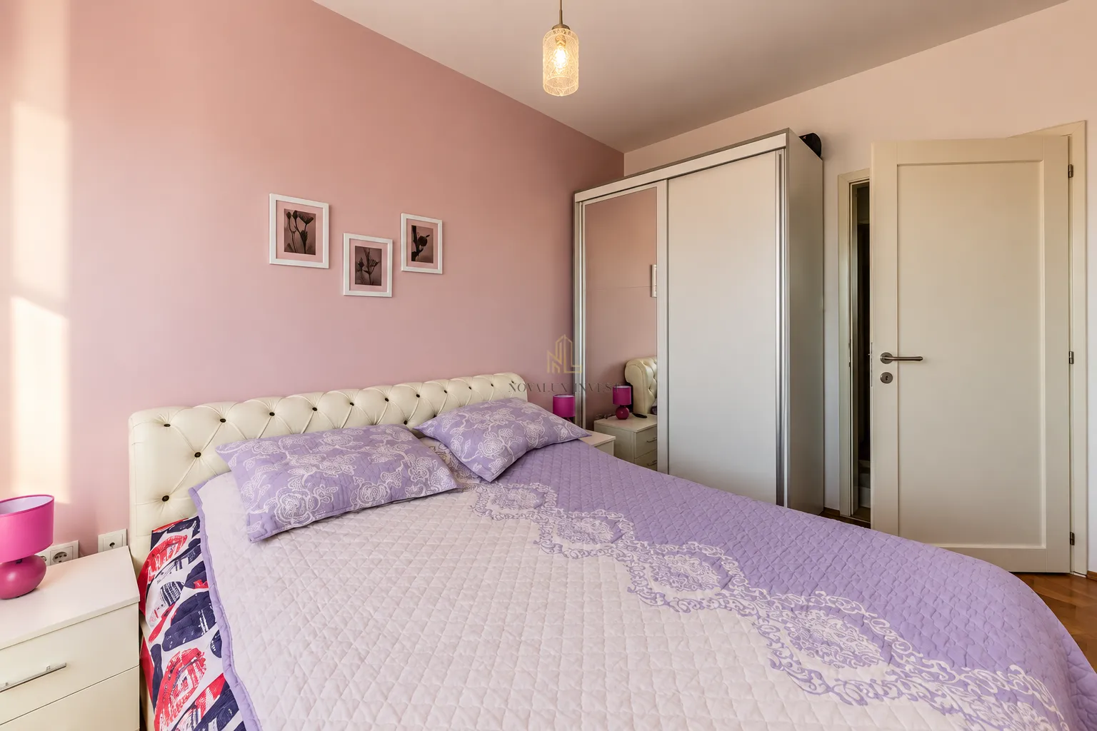Spavaća soba sa bračnim krevetom, bijelim garderoberom i roze zidom.