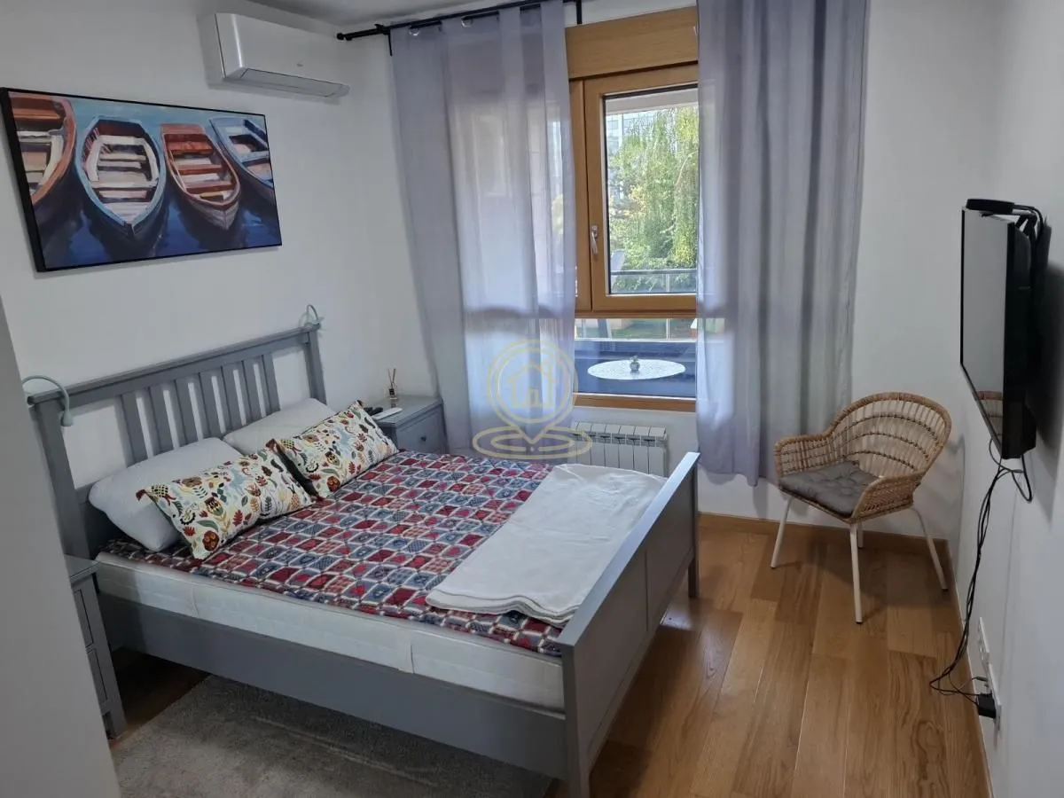 Nameštena spavaća soba sa bračnim krevetom, TV-om i prozorom sa zavesama.