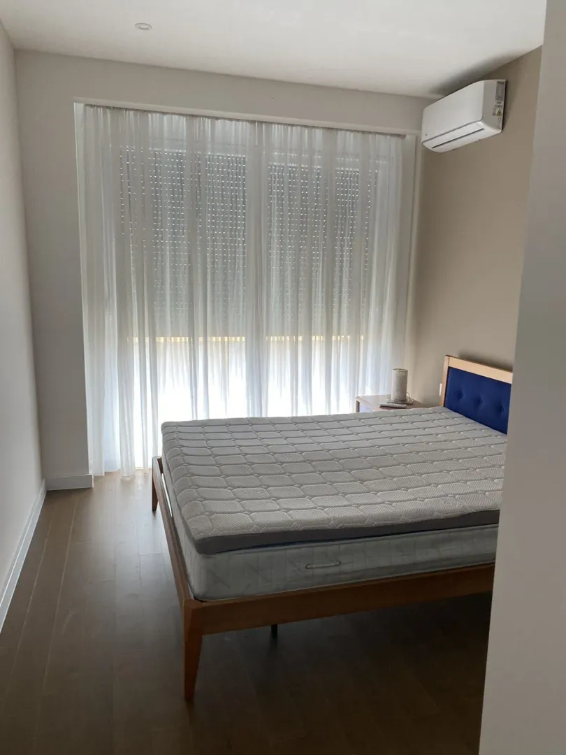 Spavaća soba sa drvenim krevetom i velikim prozorom sa bijelim zavjesama.
