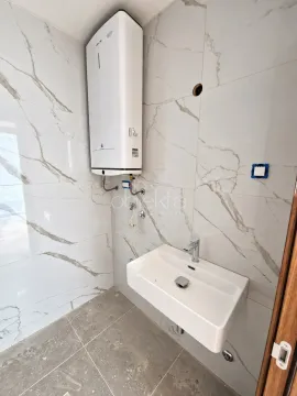 Kupatilo sa bijelim umivaonikom i bojlerom na zidu obloženom pločicama sa efektom mermera. - image 7