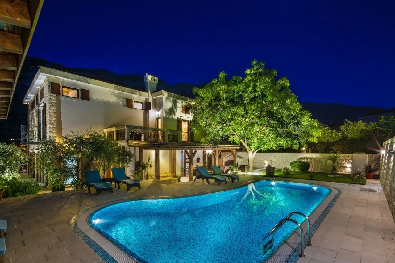 Courtyard with an illuminated swimming pool and loungers in front of a stone house at night.