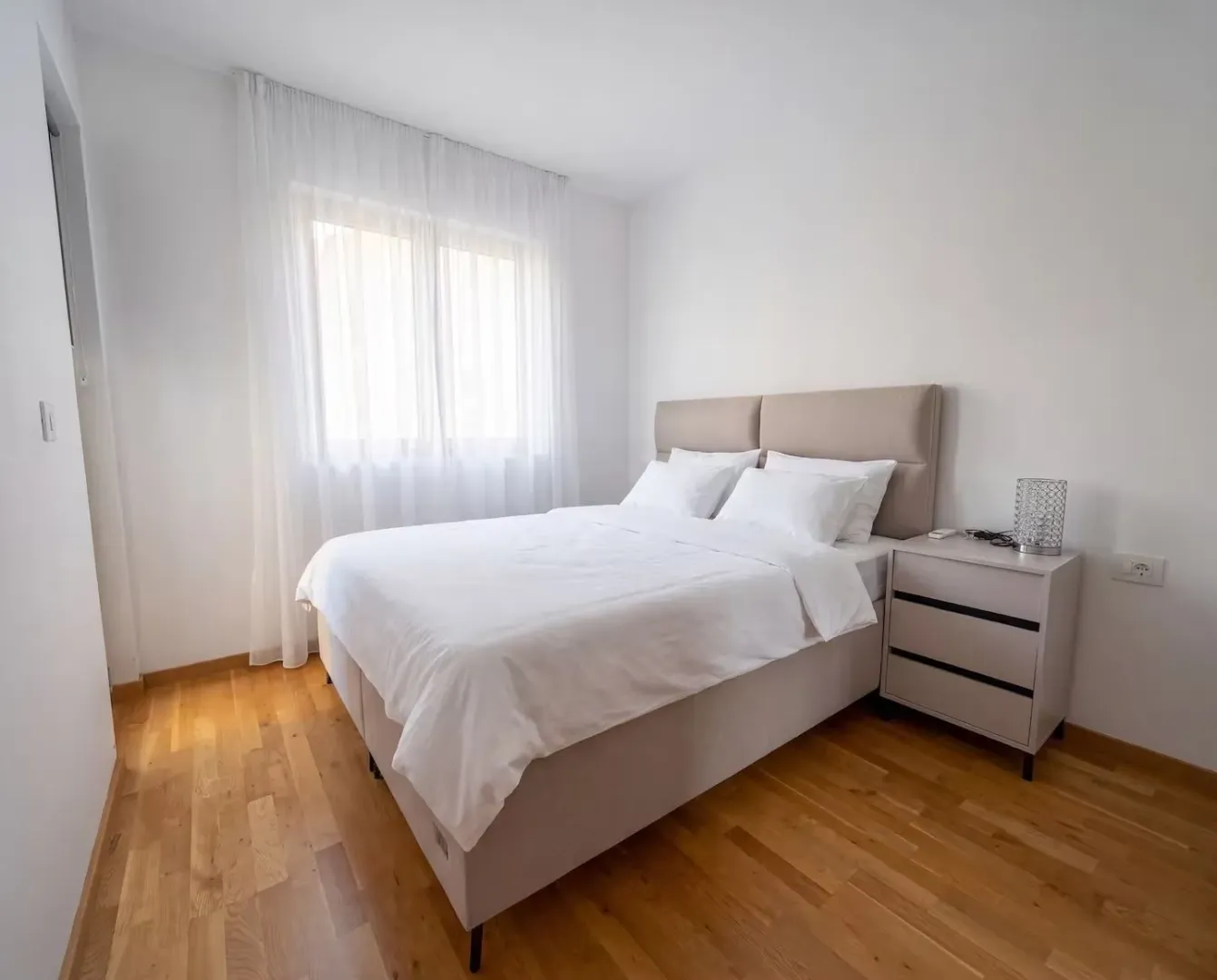 Bright bedroom with a double bed, wooden flooring, and a window with curtains.